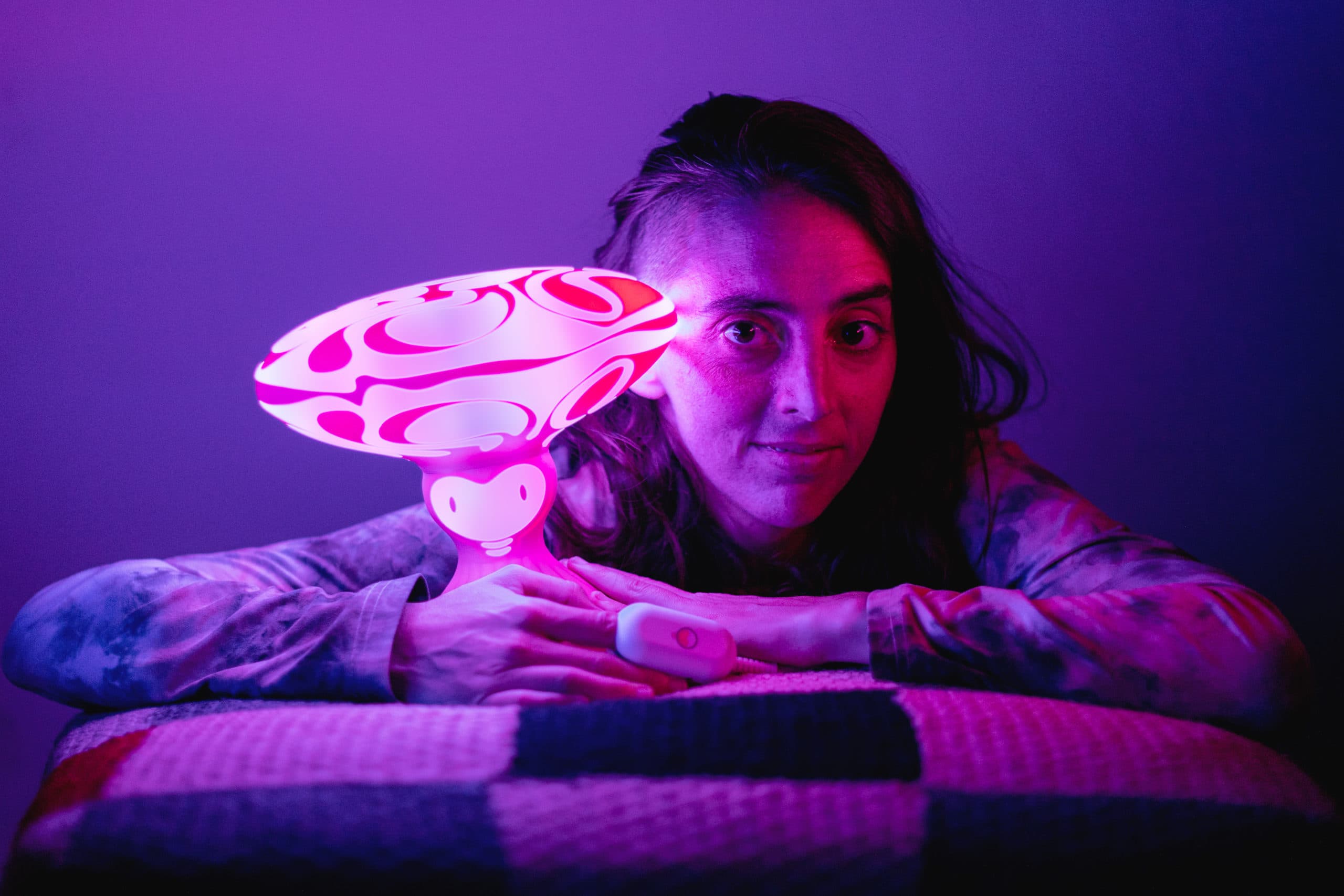 Sarit Hashkes lit in violet light beside a glowing psychedelic glass sculpture